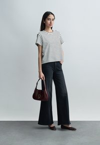Woman standing against gray wall wearing striped short-sleeve shirt, black wide-leg jeans, brown flats, and holding maroon handbag.