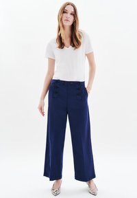 Granatowe spodnie o szerokich nogawkach z podwójnym zapięciem na guziki w talii; zestawione z białą koszulką z dekoltem w kształcie litery V i wzorzystymi butami na szpic.