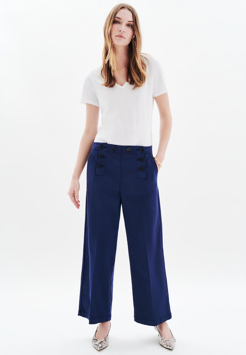 Granatowe spodnie o szerokich nogawkach z podwójnym zapięciem na guziki w talii; zestawione z białą koszulką z dekoltem w kształcie litery V i wzorzystymi butami na szpic.