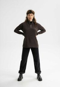 Brauner gerippter Pullover mit einem halben Reißverschluss, ausgestattet mit seitlichen Schlitzen, kombiniert mit schwarzen weiten Hosen und klobigen schwarzen Stiefeln.