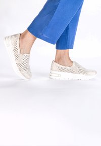 Light beige slip-on shoes with perforated design, soft texture, and a white chunky sole, worn with blue cropped pants.