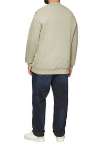 Homme portant un sweat-shirt beige, un jean bleu foncé et des baskets blanches, se tenant de dos sur un fond blanc.