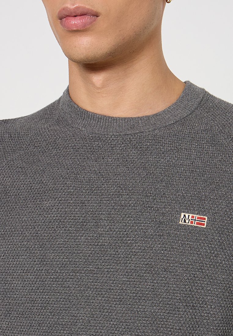 Nahaufnahme einer Person, die einen grauen strukturierten Pullover trägt, auf dessen Brust eine kleine gestickte Norwegen-Flagge und das NH-Logo zu sehen sind.