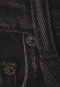 Gros plan sur un tissu en denim noir avec des coutures visibles et des rivets de couleur cuivre estampillés des lettres "WEEK DAY".
