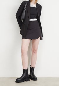 Musta räätälöity bleiseri mustan hihattoman paidan ja minihameen päällä, täydentää rouheat mustat nilkkurit. Asusteina pieni musta laukku.