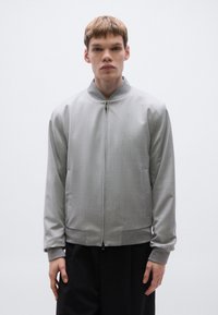 Szürke bomber dzseki enyhén texturált felülettel, bordázott mandzsettákkal és szegéllyel, cipzáras záródással és oldalzsebekkel. Simított anyag és modern kialakítás.