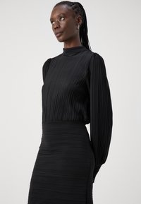 Robe noire plissée avec un col haut, de longues manches bouffantes et une silhouette ajustée. Fabriquée en tissu texturé avec un léger éclat et des plis verticaux.