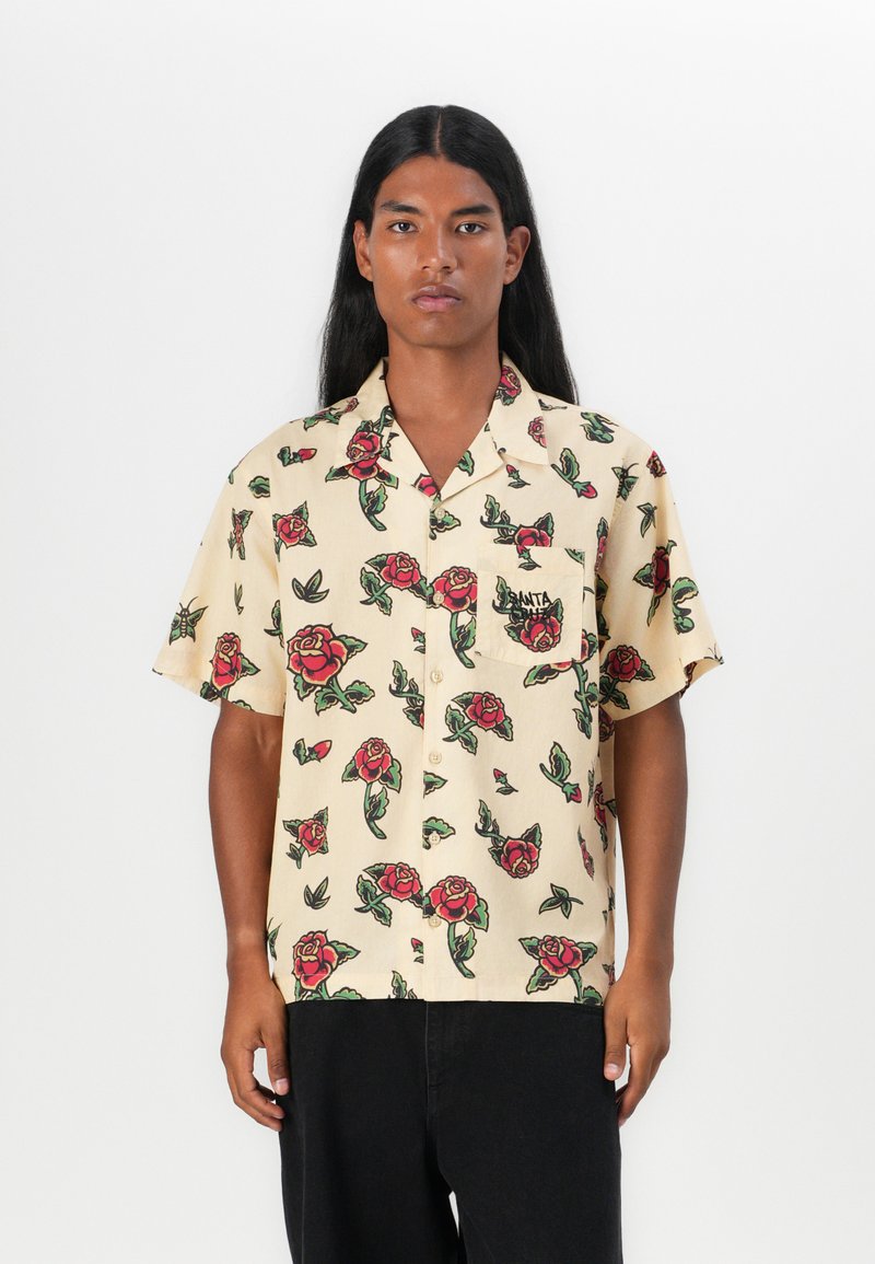 Camicia a maniche corte color beige chiaro con un motivo di rose floreali in rosso e verde. Dotata di tasca e chiusura con bottoni. Realizzata in tessuto leggero.