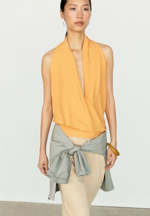 Kvinde iført ærmeløs orange draperet top, beige bukser og grå jakke bundet om taljen, stående mod ensfarvet baggrund.