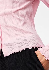 Persoană îmbrăcată într-un top roz cu mâneci lungi texturate și margini cu volane, pantaloni negri cu model ridicat, având mâna introdusă în buzunar, pe fundal alb.