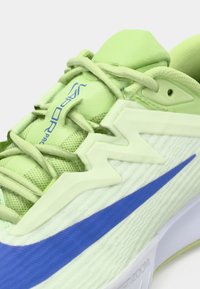 Nahaufnahme eines hellgrünen Sportschuhs mit blauem Nike-Swoosh und dem Text "Vapor Pro" auf der Zunge nahe den Schnürsenkeln.