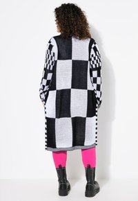 Long cardigan avec un motif à carreaux noir et blanc, texture tricotée, et des rayures noires et blanches contrastantes sur les manches et les bords.