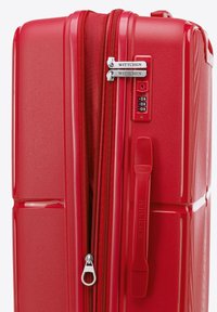 Maleta rígida roja con un acabado liso, cierre de doble cremallera, candado de combinación y nombre de marca en relieve. Las líneas verticales acentúan el diseño.