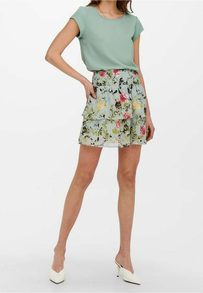 Un haut vert clair et une jupe florale à plusieurs couches de pastel bleu, ornée de motifs roses, jaunes et verts. Des mules blanches à talons hauts complètent l'ensemble.