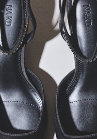 Melni augstpapēžu sandales ar siksnām, no teksturēta materiāla, ar gludu iekšējo oderi, smalku šuvumu un dekoratīvu ķēdītes akcentu.