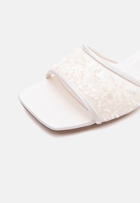 Sandale blanche à bout ouvert avec un bout carré et une large bride ornée de dentelle florale blanche et de perles décoratives.