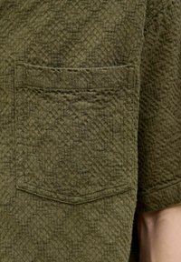Olijfgroene shirt met een gestructureerd honingraatpatroon; heeft een borstzak en korte mouwen, met een geweven stofontwerp.