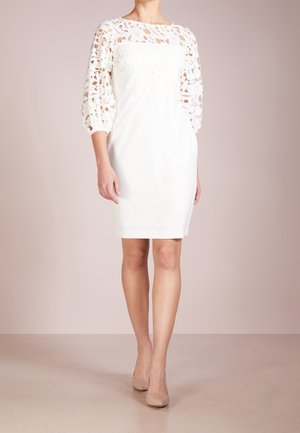 Femme portant une robe blanche mi-longue, manches en dentelle florale, et des talons hauts beige, posant devant un fond beige uni.
