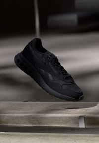 Svart atletisk sneaker med texturerad sula och snören, svävande över en konkret yta i svagt ljus.