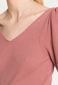 Roze geribbelde top met een V-hals en pofmouwen, met een zachte textuur en een getailleerd ontwerp. Gedetailleerde stiksels aan de randen.