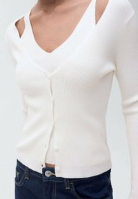 Weißer gerippter Cardigan mit V-Ausschnitt, doppellagigem Design, Knopfverschluss und langen Ärmeln, kombiniert mit dunklen Jeans.