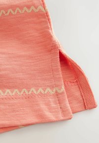 Korallfärgad textil med vävd textur, som har ett zigzagmönster i en ljusare färg längs kanten och en liten öppning vid fållen.
