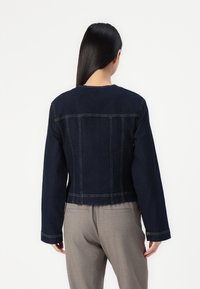 Mörkblå denimjacka med långa ärmar, kortad design, synliga sömdekorationer och en enkel rund halsringning. Bakifrån visad.