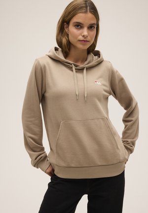 Una mujer luce una sudadera beige con capucha, un bolsillo delantero y un pequeño logotipo de FILA, combinada con pantalones negros sobre un fondo liso.