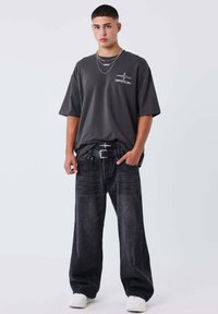 Camiseta oversized gris oscuro con logo, combinada con unos jeans de denim negros de corte holgado. El cinturón tiene una hebilla plateada. Zapatillas blancas completan el conjunto.