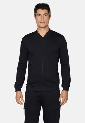 Mann trägt eine schlichte schwarze Bomberjacke mit Reißverschluss und schwarze Hose vor einem hellgrauen Hintergrund.
