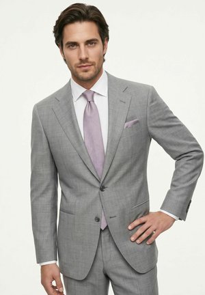 Man wearing a light grey suit with lavender tie and pocket square, standing with one hand on hip against plain background.