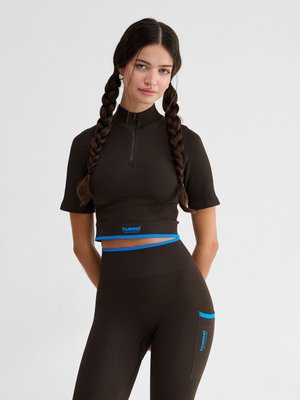 Kobieta z długimi zaplecionymi włosami, ubrana w czarny prążkowany krótki top z krótkim rękawem i dopasowane legginsy z wysokim stanem z niebieskimi akcentami odzieży sportowej Hummel.