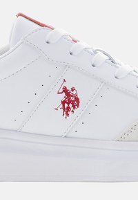 Sneaker sintetica bianca con una superficie liscia, caratterizzata da un logo di un giocatore di polo rosso, lacci bianchi piatti e piccole perforazioni sui lati.