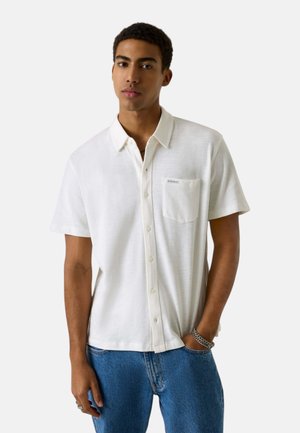 Giovane uomo che indossa una camicia bianca a maniche corte con taschino sul petto e jeans blu, in piedi con una mano in tasca.