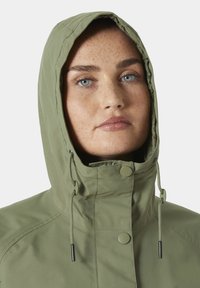 Αδιάβροχο σακάκι σε ελιά πράσινο χρώμα με κουκούλα, διαθέτει ρυθμιζόμενα κορδόνια και κλείσιμο με κουμπιά. Ομαλή υφή και ελάχιστες ραφές.