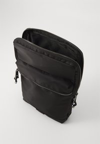 Sort nylon skuldertaske med lynlåse i toppen og frontlommen. Glat tekstur, rektangulær form og sølvfarvet lynlåsudstyr.