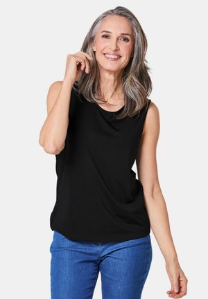 Schwarzes ärmelloses Top mit lockerer Passform und drapiertem Ausschnitt, kombiniert mit blauen Jeans. Das Model hat lange graue Haare und einen neutralen Hintergrund.