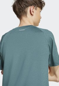 Tmavo modrozelené atletické tričko s krátkymi rukávmi, texturovaná tkanina a reflexné logo "CLIMACOOL" na zadnej časti výstrihu. Voľný strih.