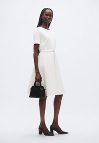 Robe blanche texturée à manches courtes avec une ceinture à la taille. Le modèle tient un sac à main noir et porte des talons pointus noirs, debout sur un fond clair.