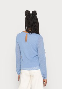 Blusa blu chiaro con maniche lunghe trasparenti, motivo a pois testurizzato, dettaglio a chiave nella parte posteriore e vestibilità comoda. Tessuto morbido e fluido.