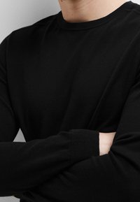 Chemise noire à manches longues en tissu tricoté doux. Présente un col rond et des poignets côtelés. La texture semble lisse et uniforme.