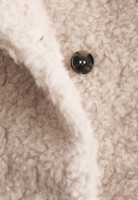 Tejido beige claro texturizado con un acabado esponjoso, que presenta un detalle de botón negro y una apariencia suave similar a la lana.