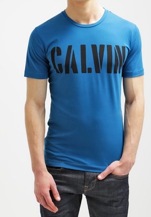 Blauw katoenen T-shirt met een ronde hals en een grote zwarte "CALVIN" print over de borst, gecombineerd met donkere spijkerbroek.