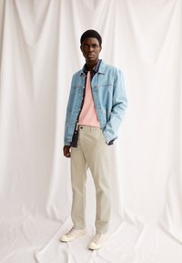 Jeansjack, roze pullover, geruit shirt, beige broek en crèmekleurige sneakers. Gladde texturen en gevarieerde kleuren creëren een gelaagde look.