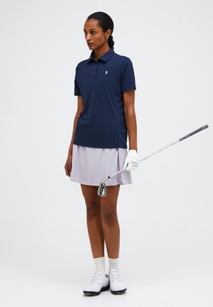 Femme debout tenant un club de golf, portant un polo bleu marine, une jupe gris clair, un gant de golf blanc, des chaussettes et des chaussures sur un fond uni.