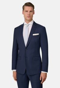 Homme portant un costume bleu marine sur mesure avec une chemise blanche, une cravate gris clair et un mouchoir blanc dans la poche, debout devant un fond uni.