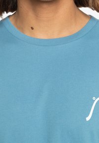 Light blue cotton t-shirt with a ribbed crew neck, featuring a small white logo on the front. Smooth texture, no visible patterns.