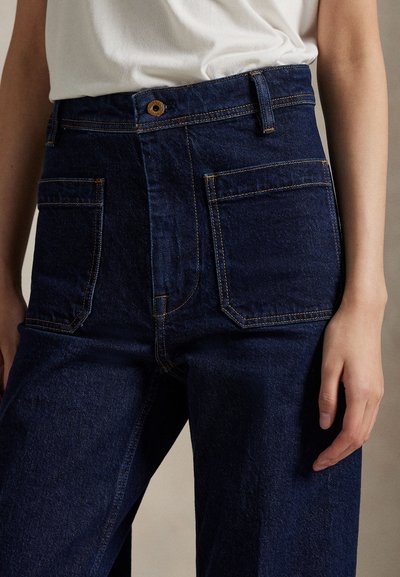 Tamsiai mėlynos, aukštos liemens džinsinės kelnės su didelėmis priekinėmis kišenėmis, dėvimos su įkištu baltu viršumi, matyti dalis liemens ir rankų.