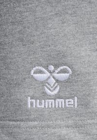 Pilkas audinys su siuvinėtu logotipu, kuriame pavaizduota stilizuota bitė ir žodis "hummel" balta spalva, pabrėžia švelnų tekstūrą.