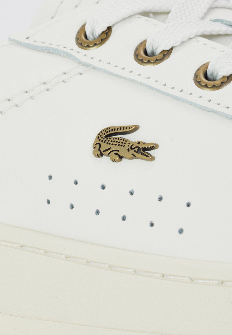 Baskets en cuir blanc avec perforations, dotées d'un logo crocodile doré et de lacets blancs. Œillets à accent en laiton et semelle texturée.